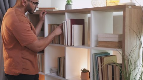 Book lover. Home library. Reading education. Intelligent man in glasses picking literature to read from bookshelf at modern living room interior.