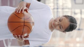 Vertical video. Poprtrait asian schoolgirl standing on basketball court holding ball raise head and smiling on the camera. Sport, education and people concept. Close up