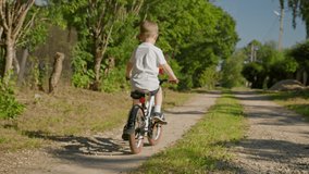 Child riding bicycle on dirt road in country village in summer sunny weather. Child rides bicycle in summer weather with toy multi-colored windmill. shooting from behind, slow motion video. Go Everyw