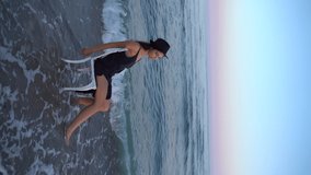 beautiful young brunette in black hat. sits on white chair by the sea. Creative romantic moody slow motion video portrait. Nostalgia moment. Black summer cocktail dress. waves touch bare feet
