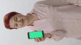 Redhead woman showing a smartphone with green screen on white background. Gamer woman pointing at smartphone with green screen, showing thumbs up with fingers.Video for the vertical story.