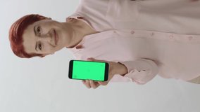 Redhead woman showing a smartphone with green screen on white background. Gamer woman pointing at smartphone with green screen, showing thumbs up with fingers.Video for the vertical story.