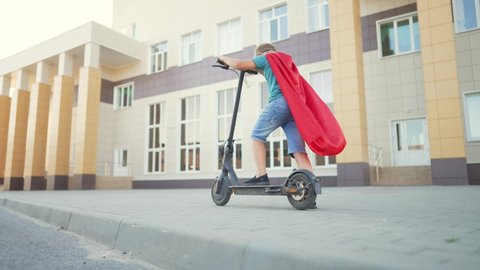 Child kid in superhero costume rides an electric scooter at school.Kid dream of travel and freedom.Schoolboy on an electric scooter in school yard.Child play in superhero costume and mask.Child winner