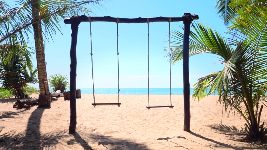 Wooden empty swings on the sandy beach with palm trees, turquoise sea and blue sky of tropical island on background. Concept of summer vacation, relax and leisure. | Shutterstock HD Video #1092565439