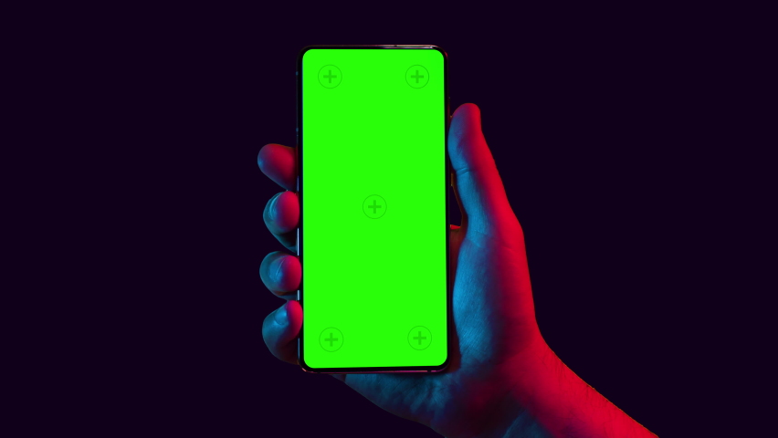 Mobile phone in hand. Holding smartphone with red blue neon lighting on dark background. Green chromakey screen with markers. Hand lifts phone up and holds it still.