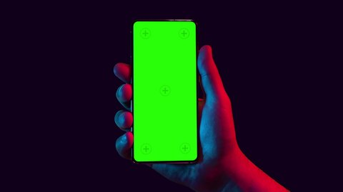 Mobile phone in hand. Holding smartphone with red blue neon lighting on dark background. Green chromakey screen with markers. Hand lifts phone up and holds it still. 库存视频