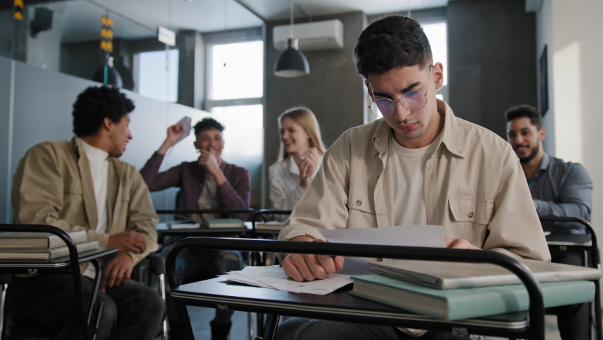 Sad worried upset disappointed caucasian student nerd wearing glasses sits in classroom at desk alone prepares for lesson reads notes. aggressive angry classmates gossiping laughing bullying poor guy Royalty-Free Stock Footage #1092568059