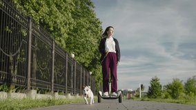 A young woman rides a hoverboard and walks her dog. Cheerful Jack Russell, runs along the sidewalk in the park - Powered by Shutterstock - Get 15% off with code: PIKWIZARD15