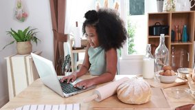 Happy african american cute small kid video call to her mother and father to show how to cook prepare healthy food  at home. African american child looking at laptop to learn bakery online alone.