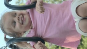 VERTICAL VIDEO: Little girl rides on gymnastic rings on open sports ground on outside. Close up portrait of cute little smiling girl swings on sports gymnastic rings in city park on sun day. Slowmo 