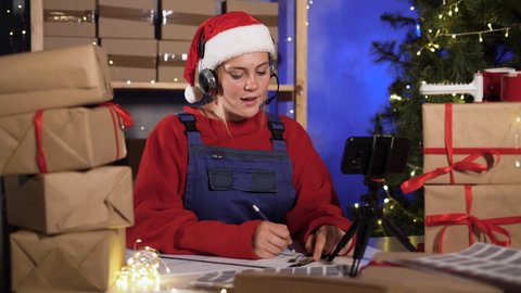 Young attractive caucasian woman dressed as santa claus takes an order online using headset and smart phone prepares delivery of goods. The concept of a big New Year or Christmas sale of goods