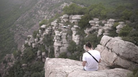 Caucasian long haired man flying a drone siting on a cliff in slow motion