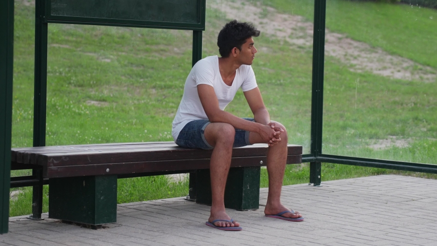 Slow motion of young Srilankan man in basic clothes and slippers casually sitting and waiting at bus stop near fields.