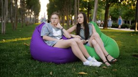 A woman and her teenage daughter are sitting together on large colored pillows on the lawn in the park.