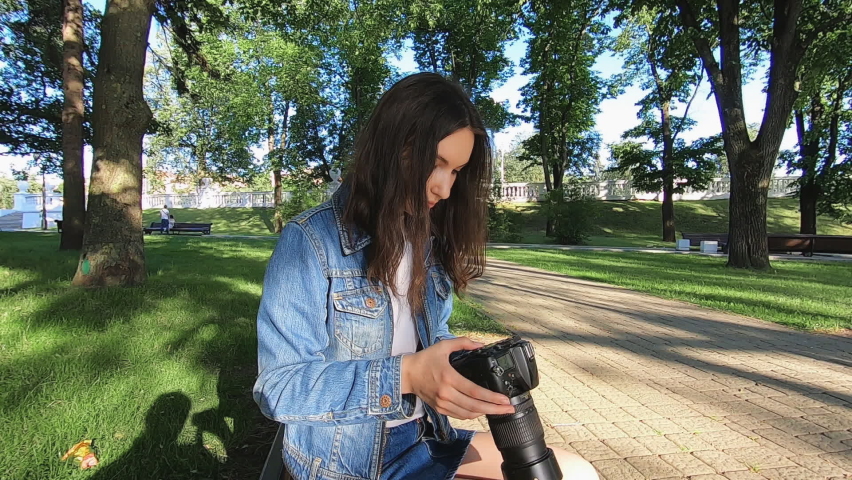 A beautiful young girl is sitting on a bench in the park looking at pictures on her camera