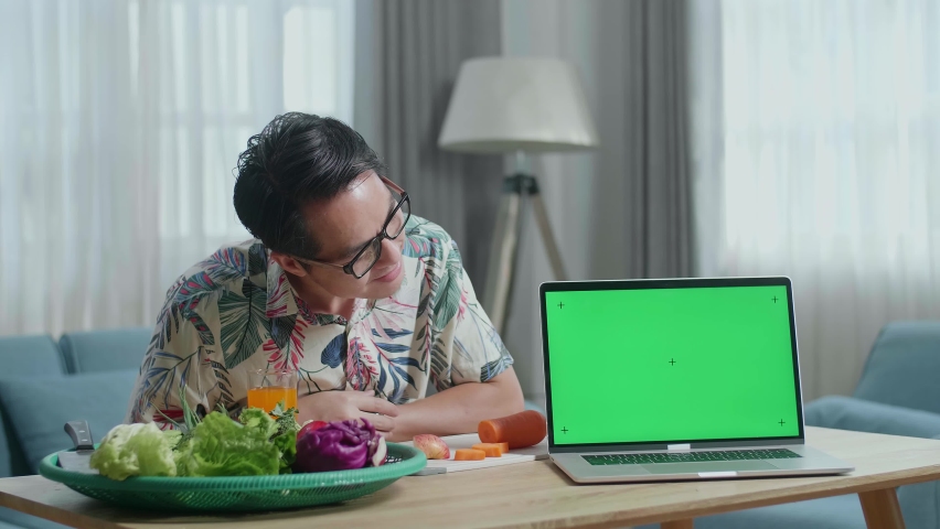 Asian Man Pointing Green Screen Laptop On The Table And Smiling To Camera While Preparing Healthy Food At Home

