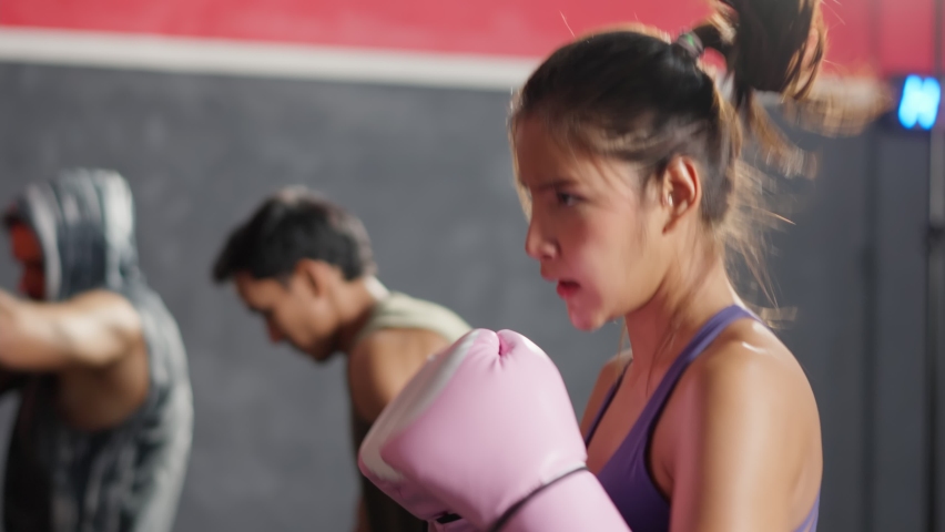 Group of athlete hitting a punching bag or sandbag at fitness gym club. Asian Fitness trainer instructor and young beautiful active sportsman and woman exercising doing boxing or Muay Thai in stadium.