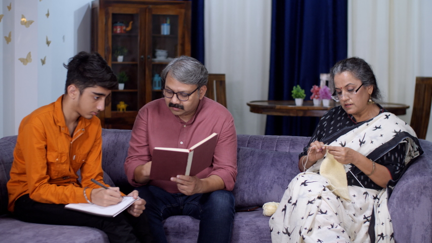 A caring father is helping his son with his studies while the mother is knitting - school assignments, hobby. Nuclear family members together in the living room - relationship and bonding, study ti...