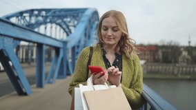 Shopping European blonde woman with sale paper bags using smart phone outdoors at city street near the bridge