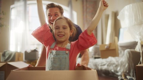 Moving in: Young Family of New Homeowners Has Fun. Father Driving Lovely Little Daughter in Cardboard Box. Cheerful Dad and Girl Racing Together Through Living Room. Tracking POV Tracking shot