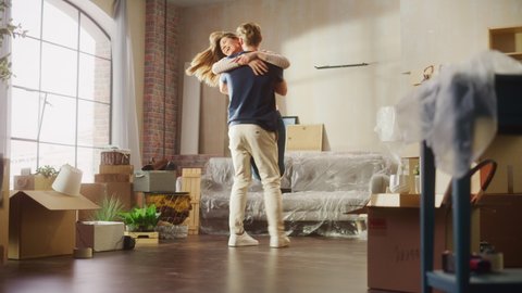 Family New Home Moving in: Happy Girlfriend Returns Home and Finds Her Happy Man Doing Unpacking, They Hug and Dance. Happy Couple in their Cozy Sunny Apartment. Mortgage Loan, Home Renovations