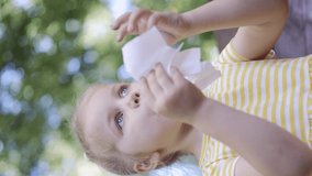 VERTICAL VIDEO: Little girl wipes her face with a paper napkin after eating, there is a milkshake on the table. Close-up of child girl sitting on park bench and wiping her face with a paper napkin.
