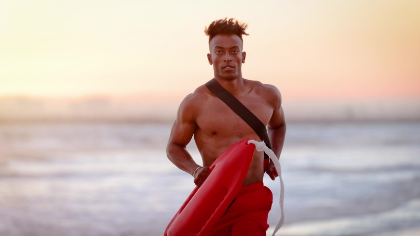 African American lifeguard running along the beach in Southern California at Sunset. Lifeguard's left hand is deformed. Royalty-Free Stock Footage #1092635439
