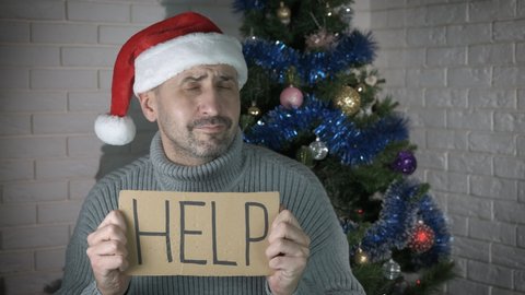 Ask for help with cardboard at holiday. A view of sad lonely man asking for help by the Christmas tree in the room.