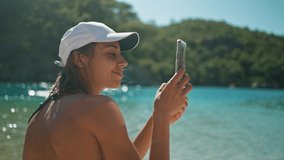 attractive young tanned woman in bikini sits on sea beach on blue water background and uses her camera on mobile phone for photo or video call. Blue Lagoon resort, Oludeniz, Turkey.