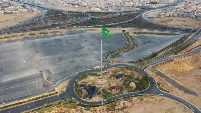 The flag of the Kingdom of Saudi Arabia flutters in the center of Riyadh at the top of the flagpole in the capital. The Kingdom Tower, Al Faisaliah Tower, and the King Abdullah Financial District Tower Royalty-Free Stock Footage #1092645077