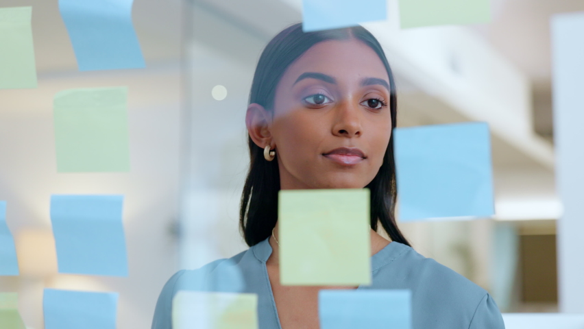 Young, thinking business woman planning information on sticky notes. A market research analyst writing a work strategy on a clear board. Thoughtful female corporate worker working in the office. Royalty-Free Stock Footage #1092656369