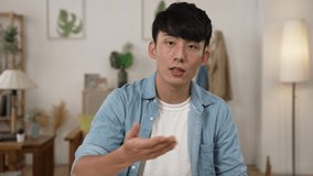 Chinese Asian male creative worker working from home looking and talking to the camera with hand gestures in an online meeting with client through video call