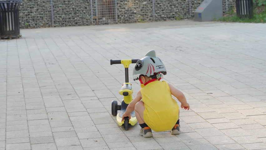 Mother teaches her toddler to ride balance bike for younger kids outdoors, 1.5 year old child with mom having fun playing with kick scooter for toddlers in the park in summer.