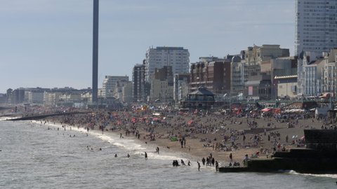 Crowded beach in Brighton UK summer with i360 and people  sunbathing and swimming in the sea.
