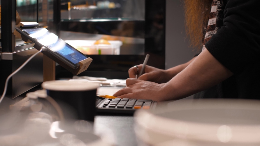Male clerk makes calculations on a calculator and writes, behind the counter in a small shop. Man makes notes on documents with a pen