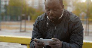 African american man sits at sport ground and types text in messenger on smartphone. Cinema 4K 60fps video