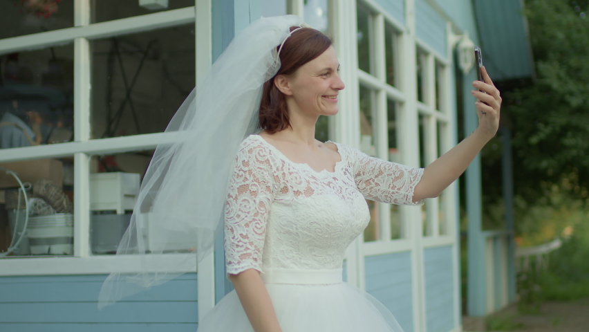 30s bride in white wedding dress using mobile phone standing near rural house. Happy newlywed holding cell phone. Slow motion, handheld camera.