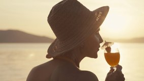 Close-up video of a woman drinking a cocktail on the beach during sunset. Girl in a hat relaxes by the ocean. High quality 4k footage