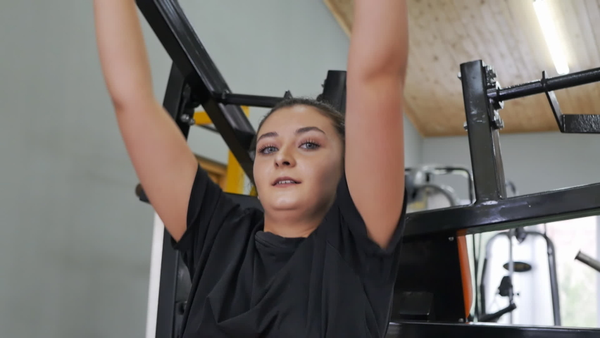 A beautiful Armenian young woman is exercising on a machine by lifting and lowering a weight with both hands. A young girl with large eyelashes of the oriental type in the fitness room Royalty-Free Stock Footage #1092688207