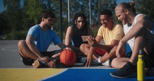 Basketball players watch videos on smartphone sitting on basketball court together. Multinational stylish sportsmen smile and laugh watching social nets closeup