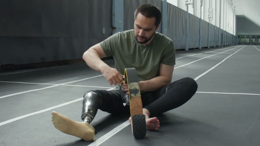 Professional sportsman with prosthetic leg sitting on floor in stadium and fixing running blade with wrench
