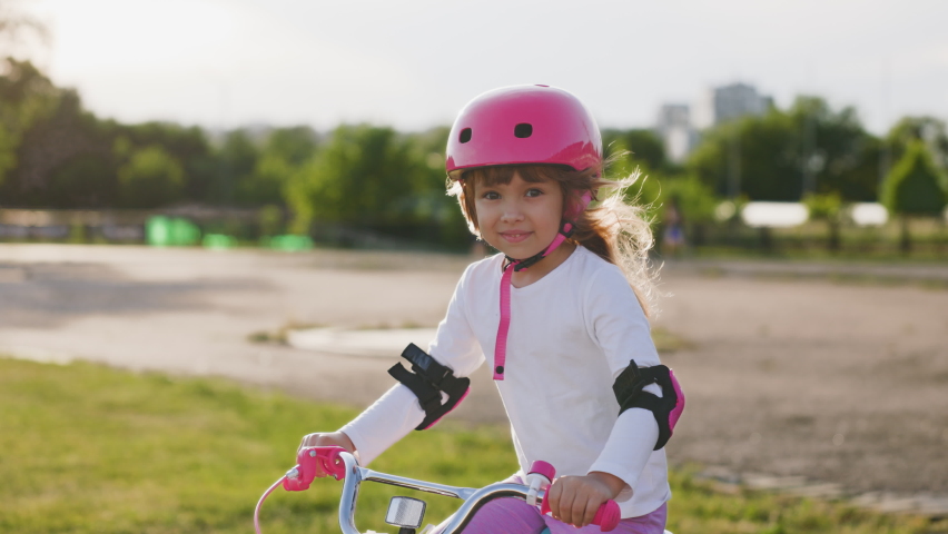 Cute light hair little girl in pink helmet in elbow and knee pads rides a bicycle smiles and feels happy. Evening sunset at stadium in background. Sport activity for children Royalty-Free Stock Footage #1092697925