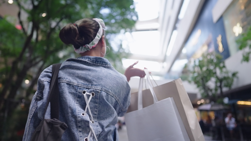 Rear View of Asian happy tourist shopper enjoys lifestyle walking in the shopping mall alone in daytime. Cheerful fashionable girl carrying paper bags leaving the shop after spending on sunny day Royalty-Free Stock Footage #1092699993