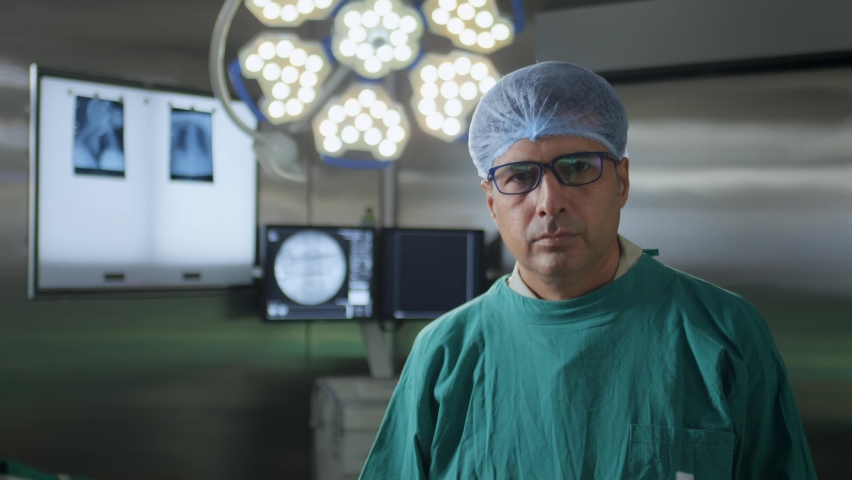 A middle-aged confident Indian Asian male surgeon, physician, or doctor wearing surgical apron, eyeglasses looking at the camera standing in an operation theatre. healthcare, medical, surgery concept  | Shutterstock HD Video #1092700017