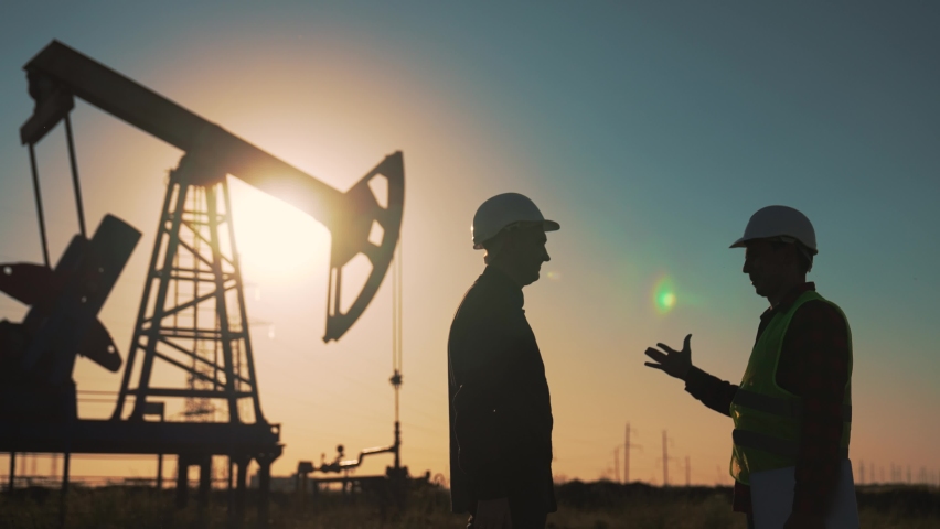 Teamwork.Oil engineer in helmet work with an oil pump.Silhouette of worker in field.Oil pump rig work at sunset.Two businessmen sign contract with handshake.Oil business concept.Work mineral deposit Royalty-Free Stock Footage #1092710331