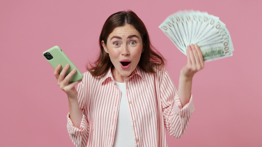 Young excited joyful caucasian woman 20s she wearing shirt t-shirt using mobile cell phone hold win fan of cash money in dollar banknotes isolated on plain pastel light pink background studio portrait | Shutterstock HD Video #1092722177