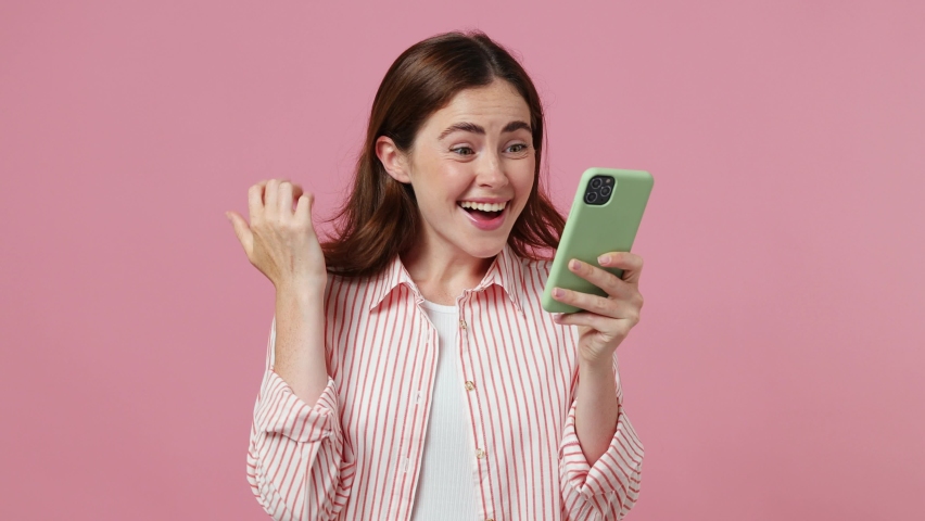 Young happy excited joyful woman she wear shirt t-shirt hold use mobile cell phone typing say wow yes just found out great big win news do winner gesture isolated on plain pastel light pink background