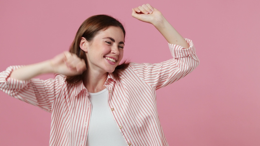 Young woman 20s she wearing shirt t-shirt dance clenching fists waving rising expressive gesticulating hands fooling around have fun enjoy celebrate play isolated on plain pastel light pink background