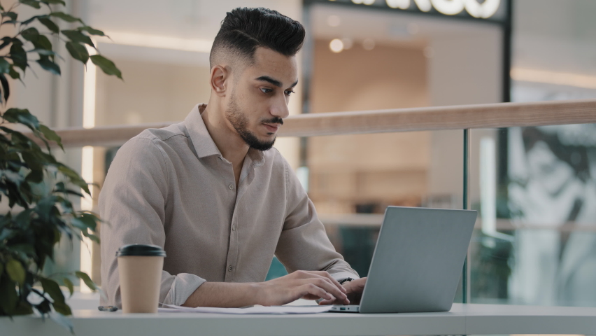 Serious busy multitasking Arabian  businessman checking corporate paperwork sitting at office cafe desk. Male worried entrepreneur working with documents analyzing financial papers with laptop Royalty-Free Stock Footage #1092722315