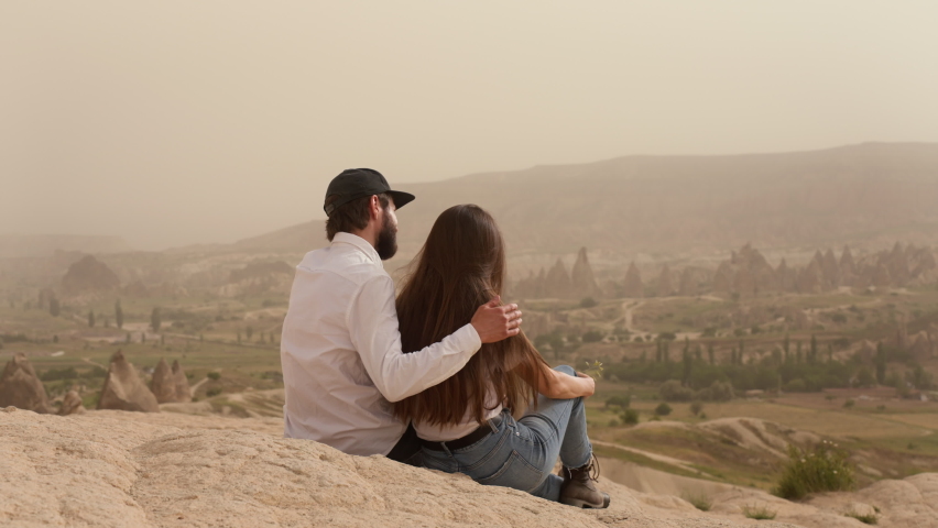 Couple of man and woman sitting on mountain cliff while hugging and admiring picturesque valley scenery with rocky formations during trip in Cappadocia on summer day. Back view 4k footage.